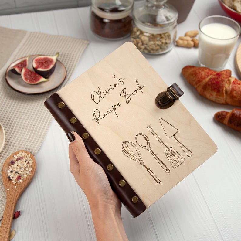 May include: A wooden recipe book with a brown leather spine and metal accents. The cover is engraved with the text "Olivia's Recipe Book" and features a drawing of kitchen utensils.