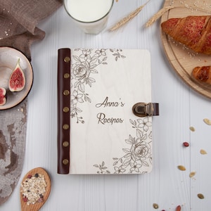 May include: A white wooden recipe book with a brown leather spine and a floral design. The book has a brass clasp and the words "Anna's Recipes" are engraved on the front cover.
