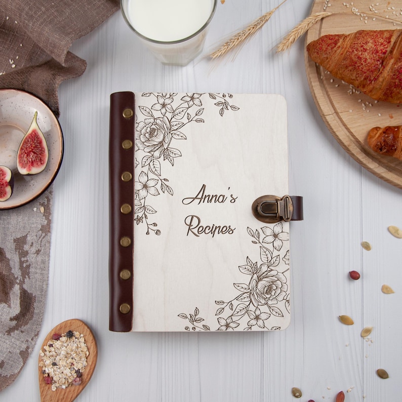 May include: A white wooden recipe book with a brown leather spine and a floral design. The book has a brass clasp and the words "Anna's Recipes" are engraved on the front cover.