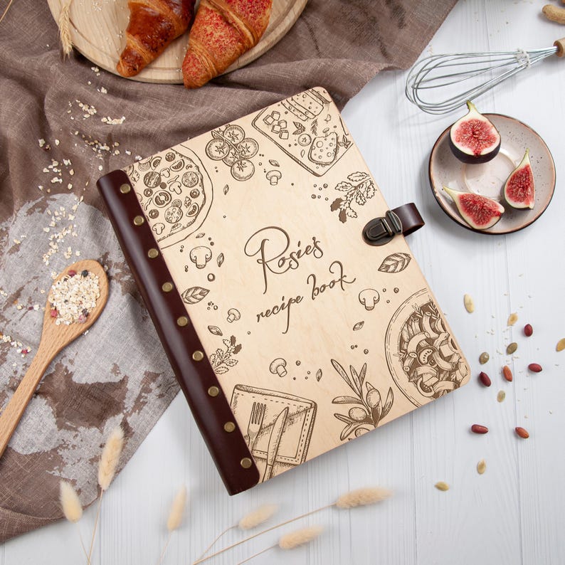 May include: A wooden recipe book with a brown leather cover and a personalised inscription that reads "Rosie's recipe book". The cover is decorated with illustrations of food, including pizza, pasta, and olives.