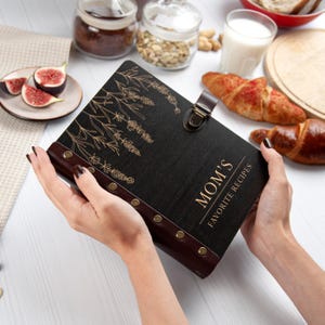 Puede incluir: Un libro de recetas encuadernado en cuero negro con letras doradas que dicen "MOM'S FAVORITE RECIPES". El libro tiene un dise&ntilde;o floral en la portada y est&aacute; sostenido por dos manos.