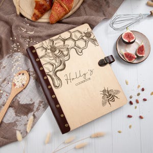 Puede incluir: Un libro de cocina de madera con una cubierta de cuero marr&oacute;n y un dise&ntilde;o de panal. La cubierta est&aacute; grabada con el texto "Holly's Cookbook" y presenta ilustraciones de abejas y panales.