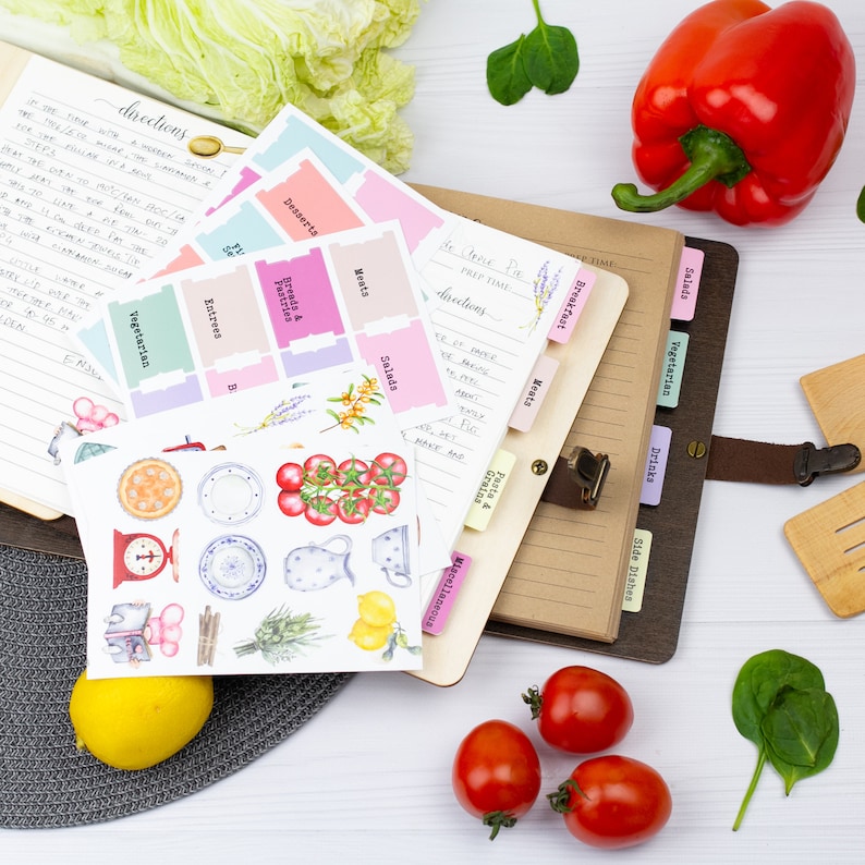 Pu&ograve; includere: Un ricettario rilegato in pelle marrone con linguette colorate per diverse categorie di pasti, come colazione, insalate e vegetariani. Il libro &egrave; aperto a una pagina con istruzioni scritte a mano e un elenco di ingredienti. Ci sono anche adesivi con illustrazioni di cibo e utensili da cucina.