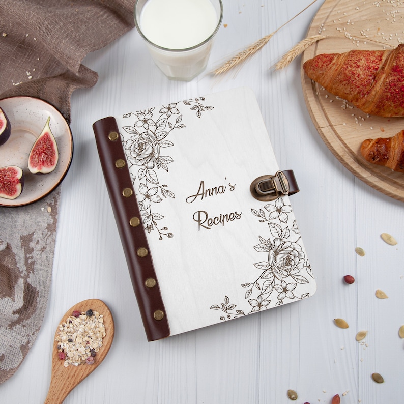 Puede incluir: Un libro de recetas blanco con una encuadernaci&oacute;n de cuero marr&oacute;n y un dise&ntilde;o floral. El libro tiene un cierre de lat&oacute;n y el texto "Anna's Recipes" est&aacute; impreso en la portada.