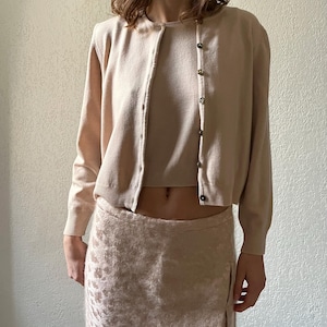 以下が含まれることがあります： ライトベージュのカーディガンと、お揃いのタンクトップとスカート。カーディガンは長袖で、ボタン留めです。スカートも同様の色で、花柄です。この服装はニュートラルカラーです。