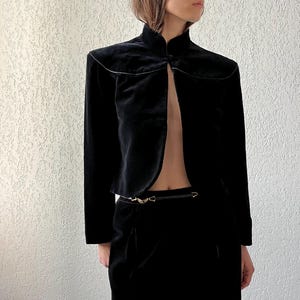 Può includere: Giacca in velluto nero con collo alto e maniche lunghe. La giacca ha un design frontale aperto unico ed è abbinata a una cintura e pantaloni neri abbinati. L'outfit è in stile vintage.