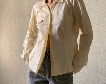 Könnte beinhalten: Ein hellgelbes Hemd mit Knöpfen und zwei Brusttaschen. Das langärmlige Hemd ist aufgeknöpft und gibt den Blick auf den Bauch frei. Das Hemd wird mit einer blauen Jeans kombiniert.