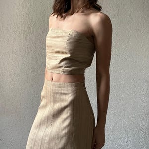 May include: A beige two-piece outfit comprising a strapless crop top and a matching skirt. The top is fitted and the skirt is high-waisted. The fabric has a subtle striped texture. The outfit is suitable for warm weather.