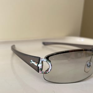以下が含まれることがあります： グラデーションレンズのリムレスサングラス。テンプルは濃いグレーで、ヒンジにはフローラルデザインが施されています。レンズはわずかに着色されており、全体的なスタイルはモダンで洗練されています。