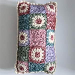 May include: A rectangular crochet pillow featuring a grid of squares in shades of dusty rose, sage green, lavender, and teal. Several squares contain a white flower motif with a rose center. The pillow is finished with a cream-colored scalloped edge.