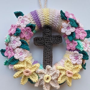 Puede incluir: Una corona de crochet con una cruz de madera en el centro. La corona está decorada con flores rosas, amarillas y verdes.