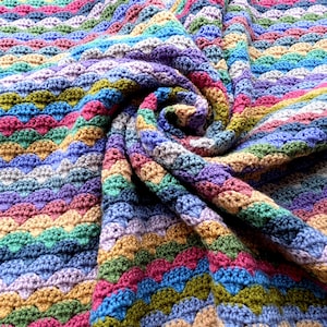 May include: A handmade crochet blanket featuring a repeating wavy shell stitch pattern. The textile displays a vibrant multicolored palette including shades of blue, green, purple, pink, and tan, arranged in horizontal stripes throughout the fabric.