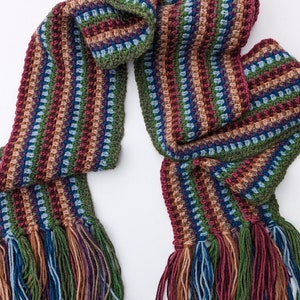 Op de afbeelding: Een gestreepte gehaakte sjaal met franjes aan beide uiteinden. De sjaal is gemaakt in verschillende kleuren, waaronder groen, blauw, bruin en rood.
