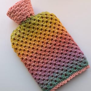 Könnte beinhalten: Eine gehäkelte Wärmflasche mit einem Regenbogen-Ombre-Muster. Die Hülle ist mit einem strukturierten Stich gearbeitet und hat einen rosa gehäkelten oberen Teil.