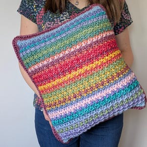 Può includere: Un cuscino all'uncinetto colorato con un motivo a righe arcobaleno. Il cuscino è realizzato con una varietà di tonalità di filato, tra cui rosa, viola, blu, verde, giallo, arancione e rosso.
