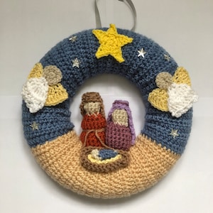 Puede incluir: Una corona de Navidad de ganchillo con fondo azul y beige. La corona presenta una estrella, dos ángeles y una escena de la Natividad con María, José y el niño Jesús.