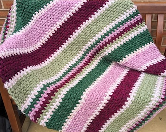 May include: A striped crocheted blanket in shades of green, pink, and burgundy. The blanket is folded and draped over a wooden bench.