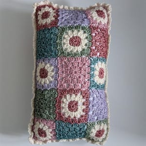 May include: Rectangular crochet pillow with a patchwork design. The pillow features squares in various pastel colors, including pink, green, and lavender, each with a central floral motif. The edges are trimmed with a cream-colored, scalloped border.