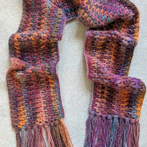 May include: A hand-crocheted scarf featuring a variegated yarn pattern in shades of deep purple, burnt orange, magenta, and hints of blue. The scarf has a dense, textured stitch pattern and is finished with long, loose fringe at both ends.