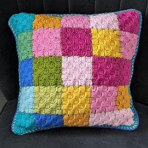 May include: A colorful crocheted pillow with a patchwork design. The pillow is made of squares in various colors, including pink, blue, green, yellow, and turquoise. The pillow has a blue and green border.