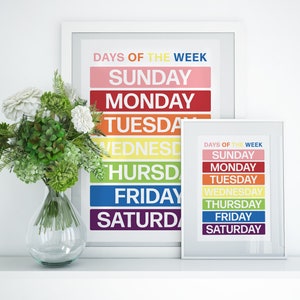May include: Two framed prints with a rainbow color scheme, each displaying the days of the week in white text. The prints are on a white shelf with a vase of white flowers and green foliage.