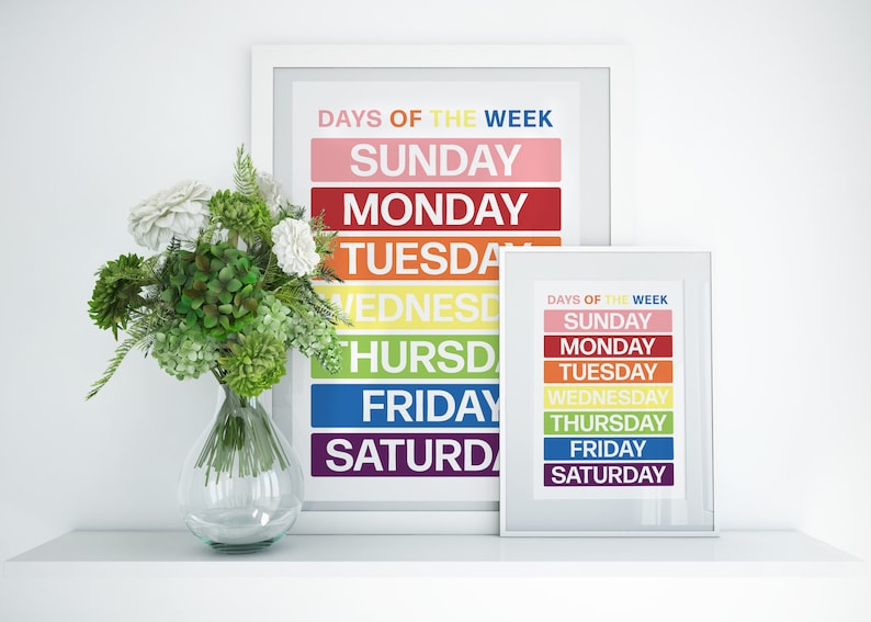May include: Two framed prints with a rainbow color scheme, each displaying the days of the week in white text. The prints are on a white shelf with a vase of white flowers and green foliage.