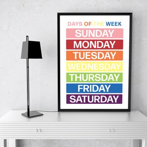 May include: A black framed poster with the text "Days of the Week" in white. The days of the week are listed in a column, each in a different color: Sunday (pink), Monday (red), Tuesday (orange), Wednesday (yellow), Thursday (green), Friday (blue), and Saturday (purple).