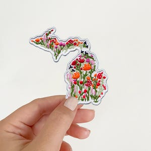 May include: An embroidered patch shaped like the state of Michigan, featuring a dense pattern of colorful wildflowers in shades of red, orange, pink, and green. The patch has a white border and is filled with detailed floral stitching throughout.