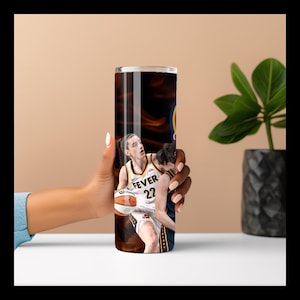 May include: A tall, cylindrical tumbler featuring a basketball player in action, wearing a white and gold jersey with the number 22 and the word "FEVER". The background has a fiery, orange and brown design. The tumbler is held by a person.