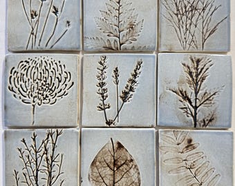 May include: A set of nine square ceramic tiles arranged in a grid, each featuring a unique dark brown botanical imprint on a light gray background. The designs include various leaves, grasses, and flower heads pressed into the smooth clay surface.