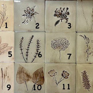 May include: A grid of twelve square ceramic tiles, each featuring a unique botanical imprint in brown ink on a light beige or pale green background. Each tile is numbered from one to twelve in black, showcasing various plant species like leaves, flowers, and grasses.