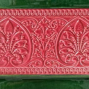 May include: A rectangular ceramic tile featuring a raised relief pattern of two symmetrical floral motifs in a deep rose pink color. The tile is bordered by a beaded edge and is surrounded by dark green tiles with intricate embossed details.