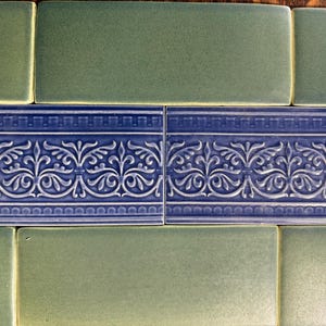 May include: A collection of rectangular ceramic tiles arranged in rows. The top and bottom rows consist of solid, matte sage green tiles. The middle row features two deep blue tiles with a raised, intricate embossed scrollwork pattern.