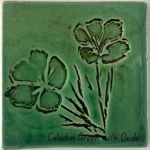May include: A square ceramic tile with a glossy celadon green glaze featuring a debossed floral design with dark oxide detailing. The text "Celadon Green with Oxide" is printed in black at the bottom of the tile surface, highlighting the finish.