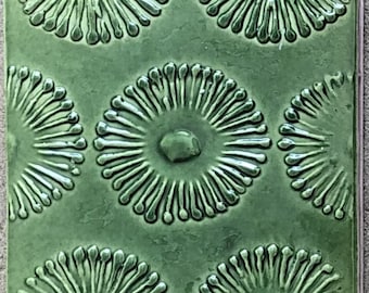 May include: A square ceramic tile with a glossy olive green finish. The surface features a repeating embossed pattern of circular, sunburst-like motifs with radiating lines and a central raised dot, creating a textured, tactile appearance.