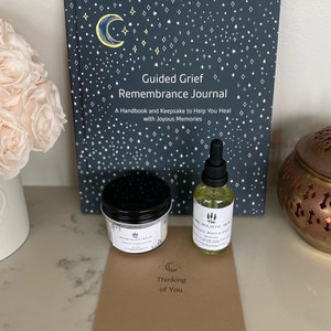 Puede incluir: Una colección de artículos de autocuidado, que incluye un diario titulado "Guided Grief Remembrance Journal", un tarro de sales de baño, una botella de aceite facial y una tarjeta que dice "Thinking of You". El diario tiene una cubierta azul oscuro.