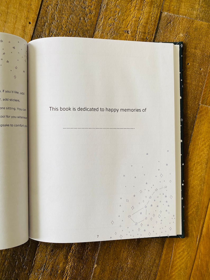May include: An open book with a white page that says "This book is dedicated to happy memories of" with a line for writing. The page is surrounded by a black page with a silver constellation design.