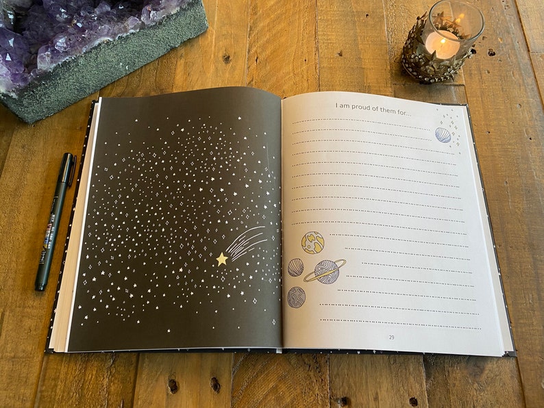 May include: An open journal with a black cover and white stars on the left page. The right page has the text "I am proud of them for..." and a drawing of planets and stars. The page number 23 is visible in the bottom right corner.