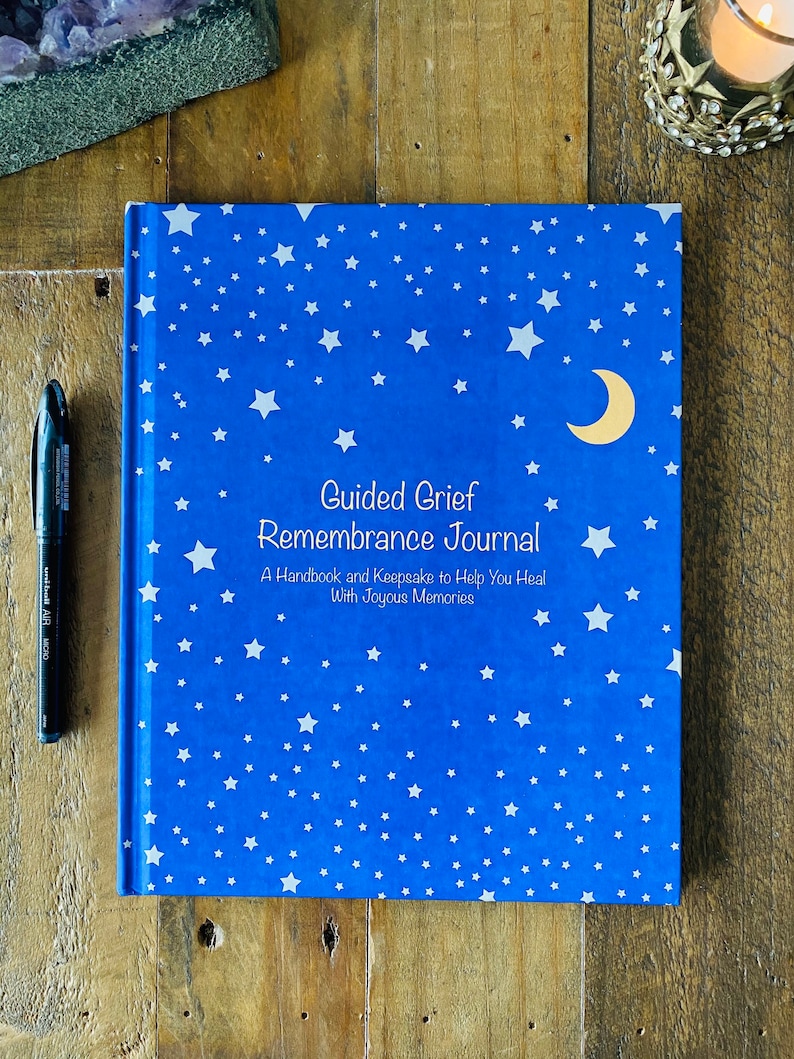 May include: A blue journal with a white star pattern and a gold crescent moon. The cover reads "Guided Grief Remembrance Journal: A Handbook and Keepsake to Help You Heal With Joyous Memories".