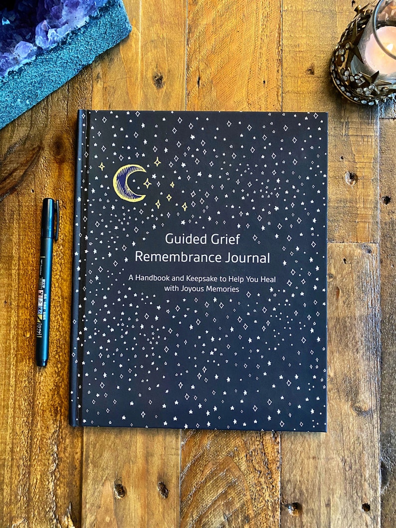 May include: A black journal with a gold crescent moon and stars design. The title is "Guided Grief Remembrance Journal" with the subtitle "A Handbook and Keepsake to Help You Heal with Joyous Memories".