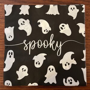 May include: A square paper napkin with a black background featuring a repeating pattern of white ghosts with two eyes and a mouth. The word spooky is printed in white cursive script across the center of the napkin.