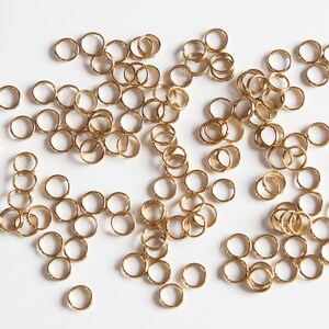 May include: A large quantity of small, gold-coloured jump rings. The rings are scattered on a white surface.