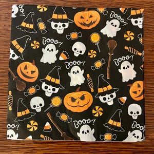 May include: A square paper napkin with a black background featuring a repeating Halloween pattern of white ghosts, orange jack-o'-lanterns, skulls wearing witch hats, brown broomsticks, colorful candies, and the white text "Booo!".