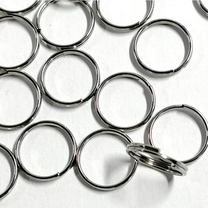 May include: A collection of silver-coloured metal split rings arranged in a scattered pattern. Each ring features a circular design with a double-loop construction, creating a durable and functional component for keychains or various organisational tasks.
