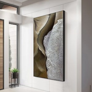 May include: Abstract wall art with a black frame. The artwork features a textured design with shades of brown and white, resembling a landscape or aerial view of a coastline. The piece is displayed on a white wall.