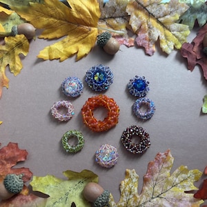 Könnte beinhalten: Eine Auswahl an geschmückten Schmuckstücken, darunter Ringe und dekorative Broschen, in verschiedenen Farben wie Blau, Orange und Lila. Der Schmuck ist mit Herbstblättern und Eicheln arrangiert, was ein saisonales Thema schafft.