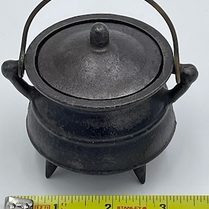 May include: A small black cast iron cauldron with a lid and a brass handle. The cauldron is about 3 inches tall and has three legs.