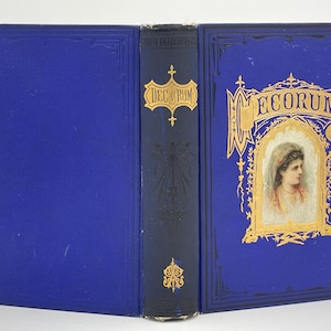 May include: A blue book with gold lettering on the spine and cover. The title of the book is "DECORUM". The cover features a gold frame with a woman's portrait inside.