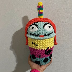 May include: A crocheted amigurumi doll shaped like a character from the movie "The Nightmare Before Christmas". The doll is wearing a red, yellow, and blue hat and has a green face with black eyes and a stitched mouth.