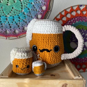 Peut inclure: Trois mugs à bière en crochet, un grand, un moyen et un petit, avec des dessus de mousse blancs et des corps orange. Les mugs ont des visages mignons avec des yeux, une moustache et un sourire. Les mugs sont posés sur une surface en bois.